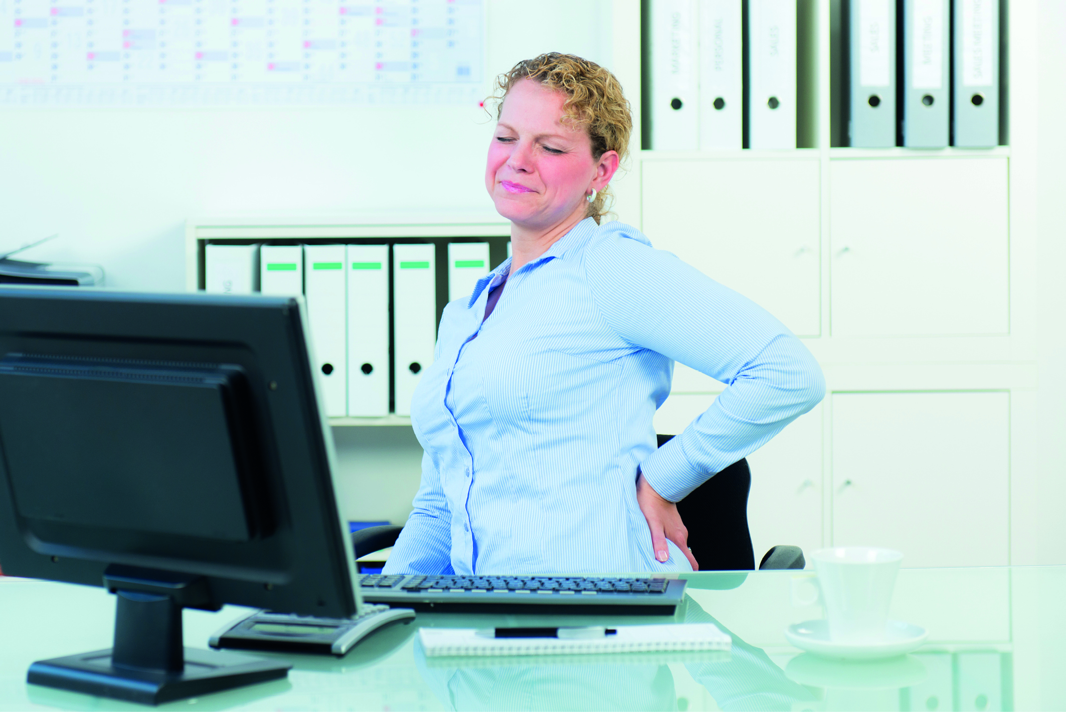 Rückenschmerzen: eine Frau sitzt an ihrem Schreibtisch und hält sich vor Schmerzen den Rücken