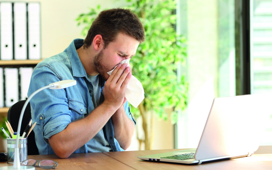 Ein Mann sitzt am Schreibtisch vor seinem Laptop und putzt sich seine Nase. Er geht trotz Krankheit ins Büro