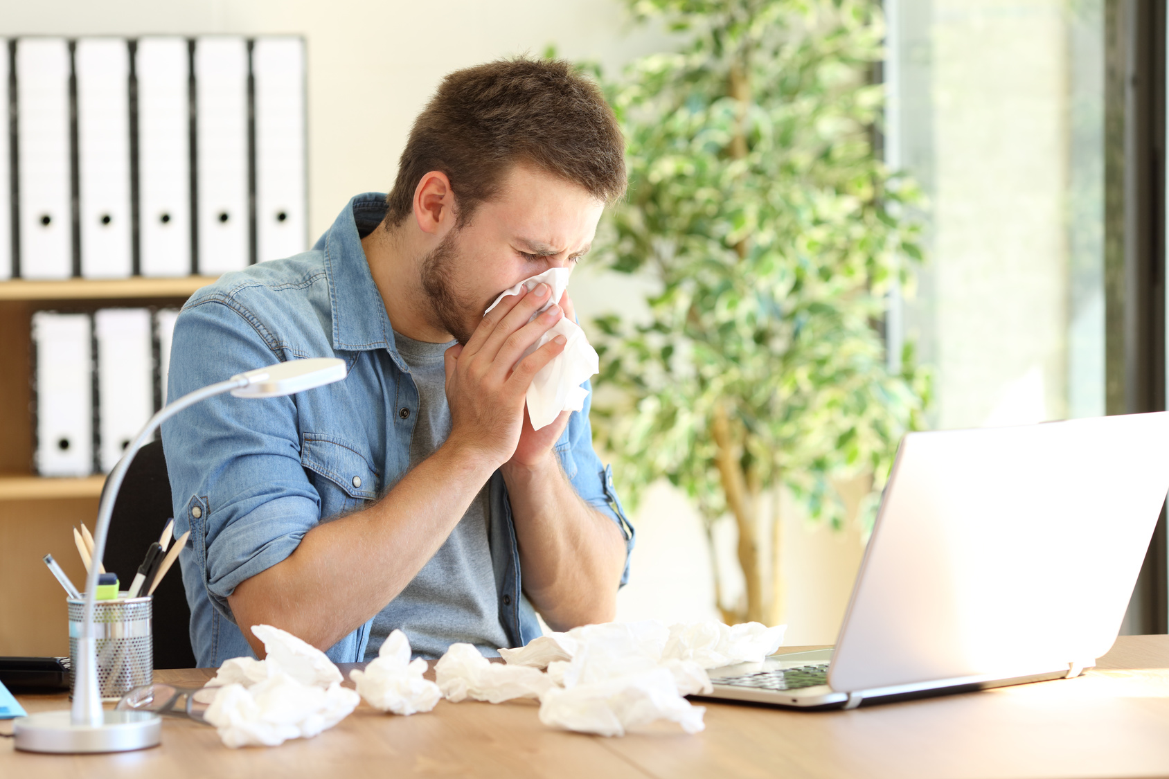 Ein erkälteter Mann sitzt im Büro vor einem Laptop und putzt sich die Nase. Neben ihm liegen viele benutzte Taschentücher.