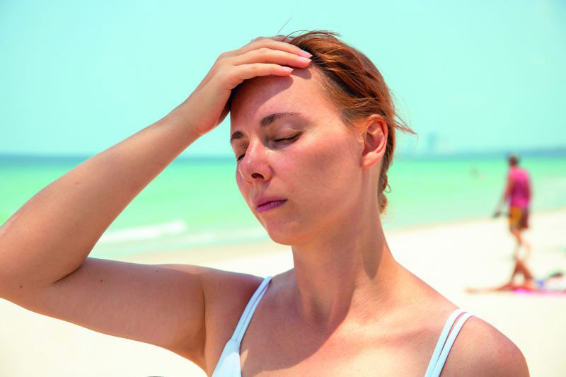 Krank im Auslandsurlaub: Eine Frau steht mit geschlossenen Augen am Strand und hat eine Hand auf die Stirn gelegt.