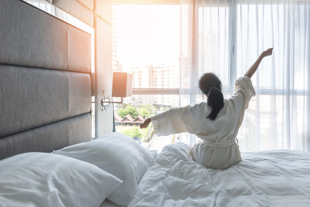 Starkes Immunsystem: eine Frau sitzt mit dem Blick in Richtung Fenster auf der Bettkante und streckt sich.