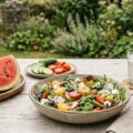 Leichte Sommerküche: bunter Salat, Wassermelone, Gurken und Tomaten auf einem Holztisch im Freien